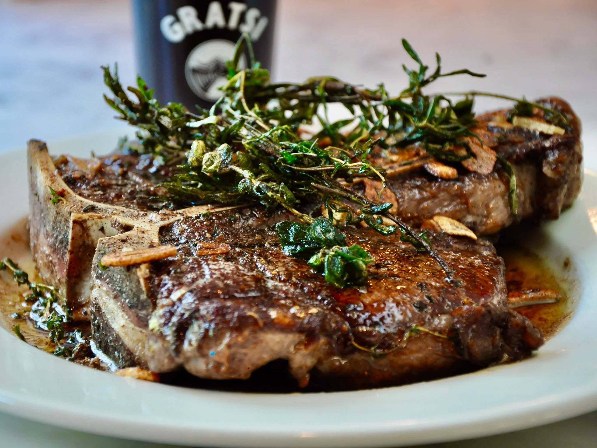 Two cooked ribeye steaks with herbs and garlic slices on top, served on a white plate. A bottle with the word GRATSI is blurred in the background.