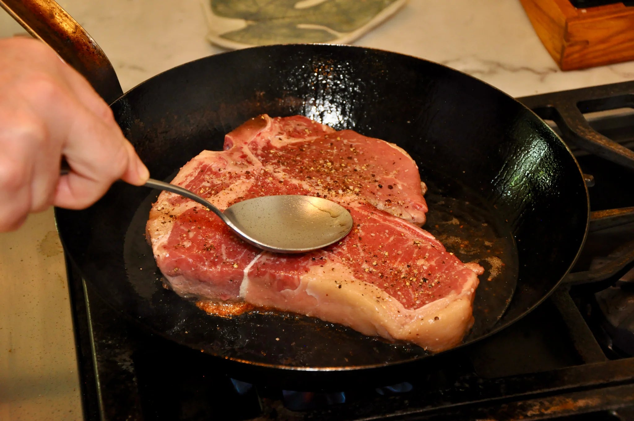A hand presses a metal spoon against a seasoned raw steak in a black skillet on a stovetop, beginning the cooking process.