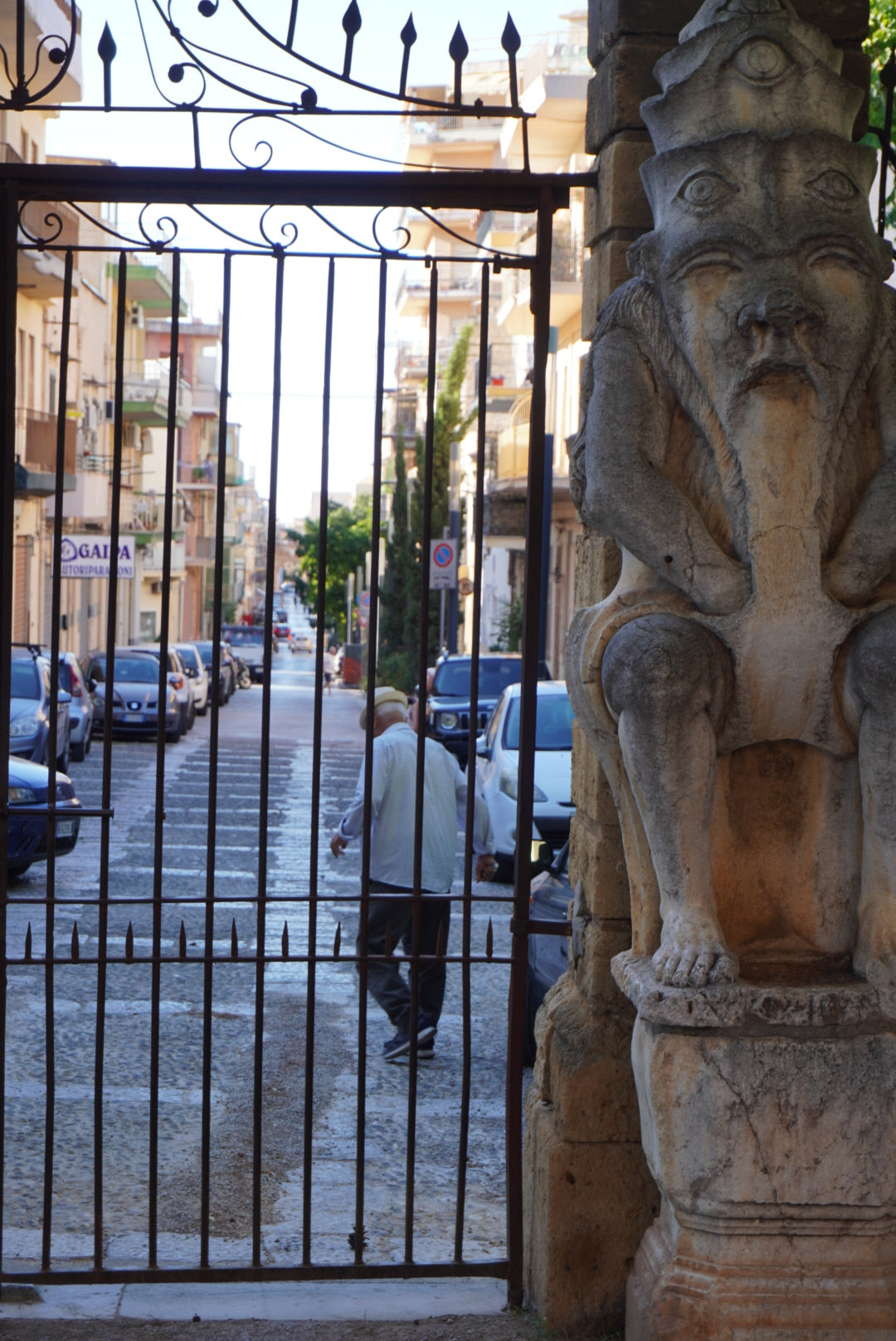An elderly man walks through an open iron gate on a city street, beside a large, weathered stone statue of a crouching figure with an expressive face. Parked cars and apartment buildings line the street.