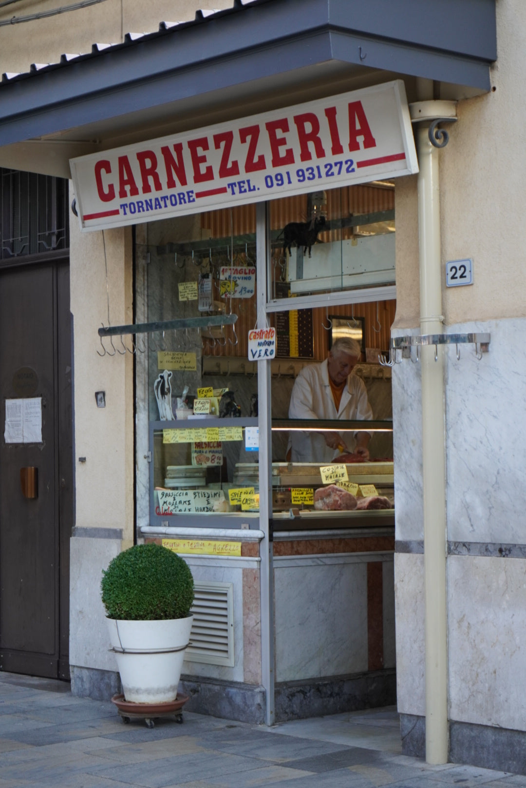 A butcher shop with a sign reading “CARNEZZERIA” above the entrance. A butcher works behind the glass display filled with meats. A potted plant sits outside next to the doorway. The shop address is 22.