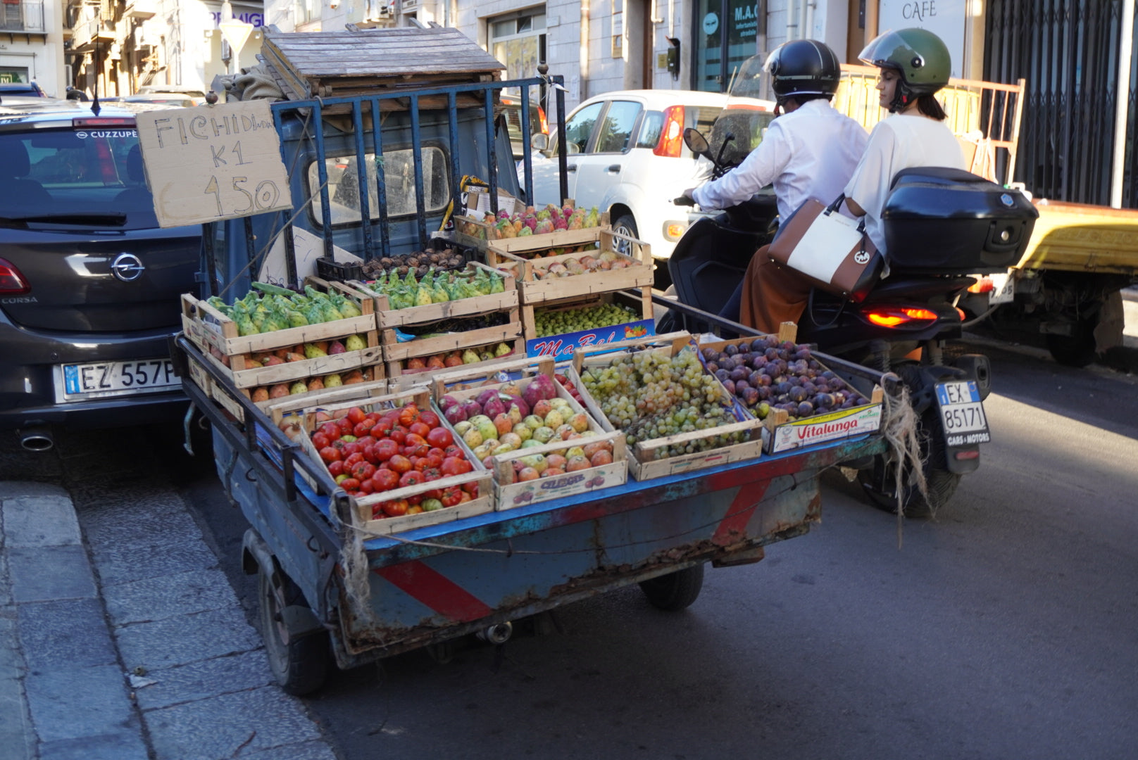A blue cart loaded with crates of fresh fruits, including prickly pears, is parked on a city street. A sign on the cart displays the price. Two people on a scooter are stopped next to the cart. Cars are parked along the street.