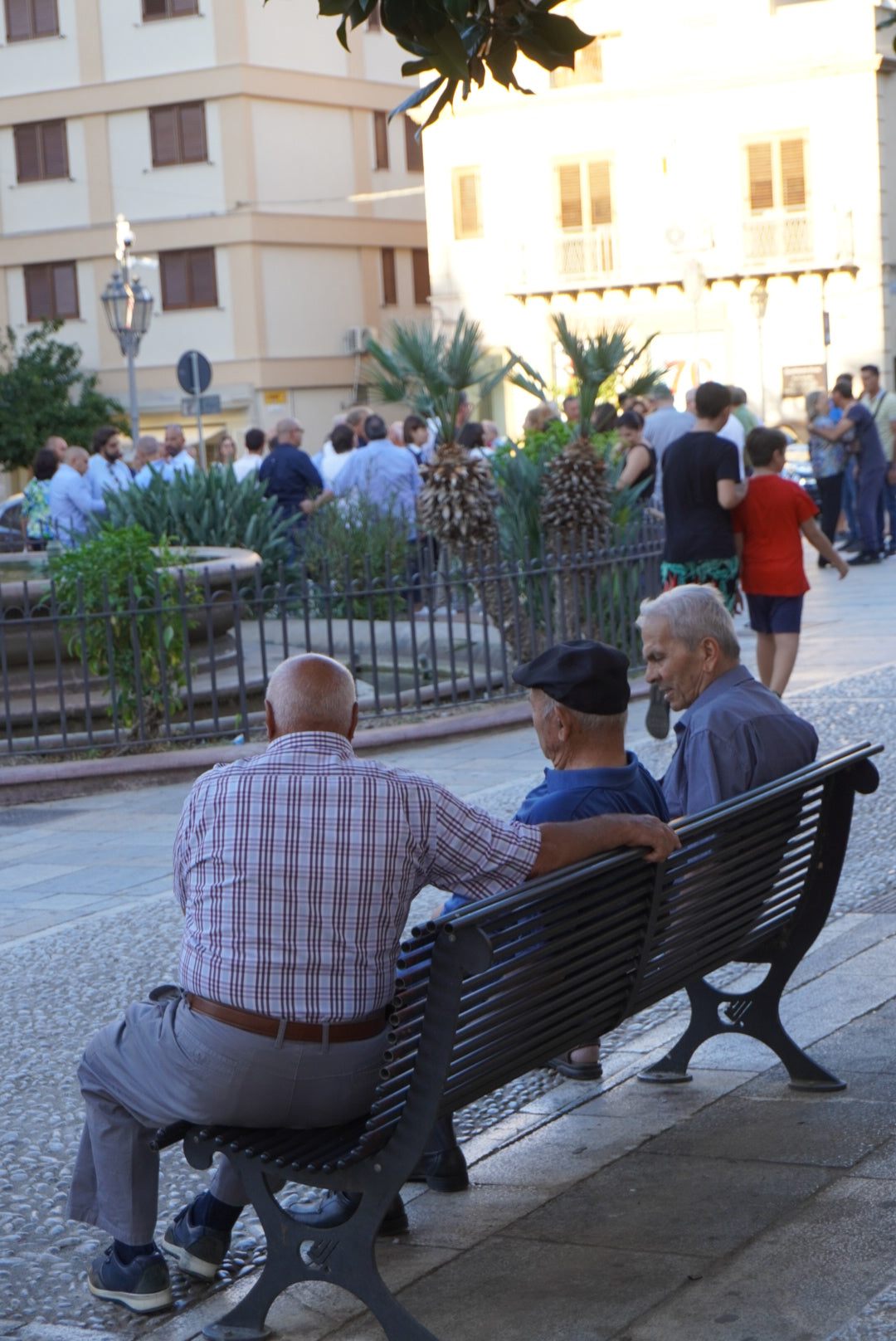 Three elderly men sit on a bench in a city square, facing a crowd gathered near a fountain and plants. Buildings and greenery are visible in the background under daylight.
