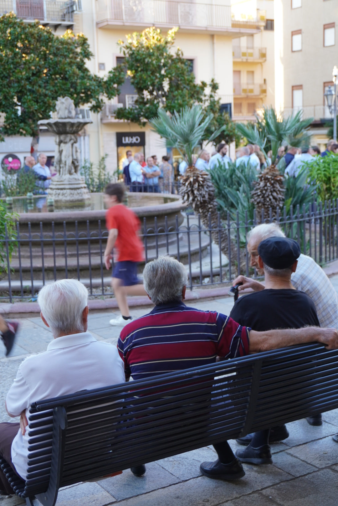 Three elderly men sit on a bench facing a fountain in a town square, while a child runs by and people gather in the background among trees and buildings.