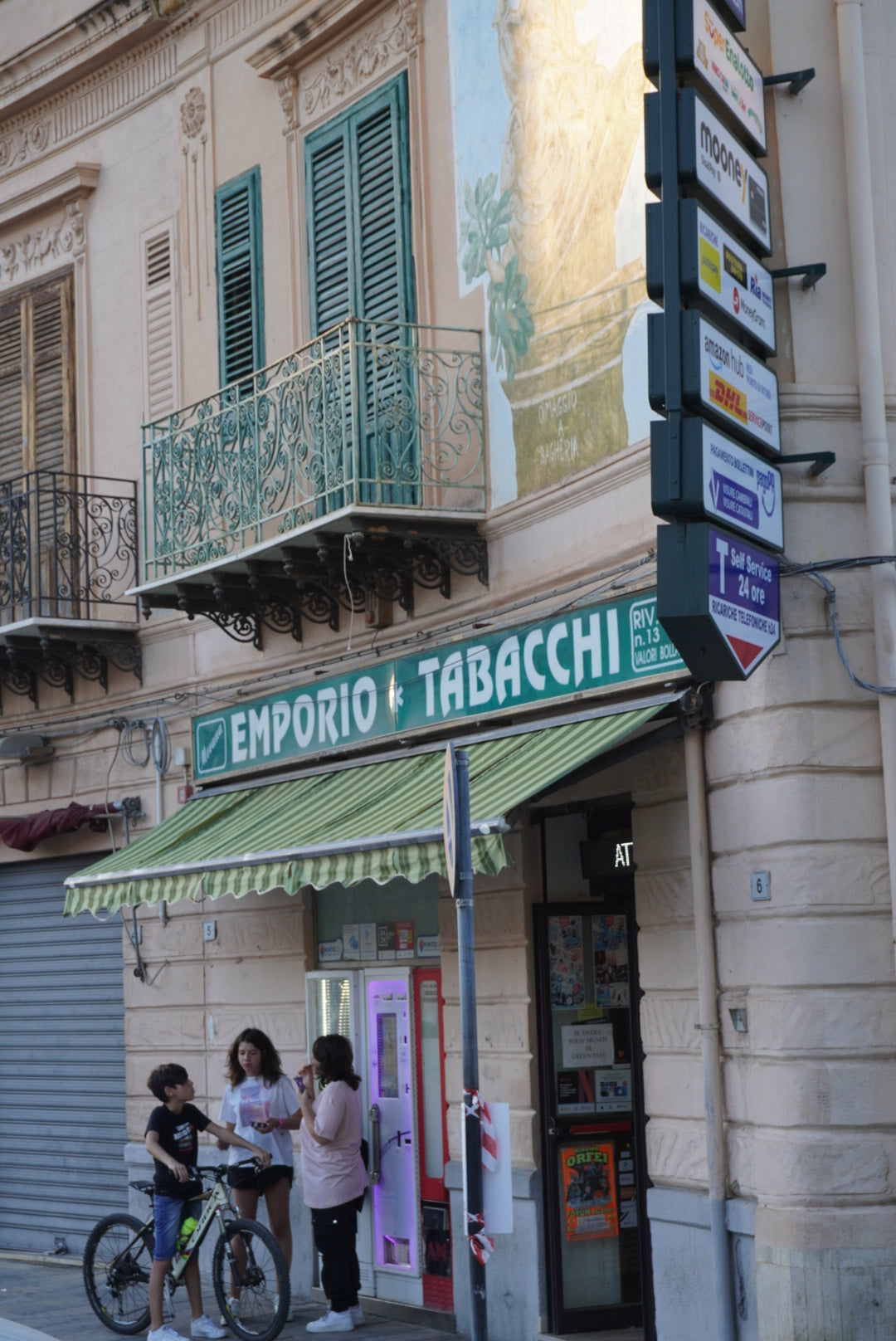Three people stand outside an Emporio Tabacchi shop on a city street, with a green-striped awning above. The building has ornate balconies and several signs are visible above the entrance.