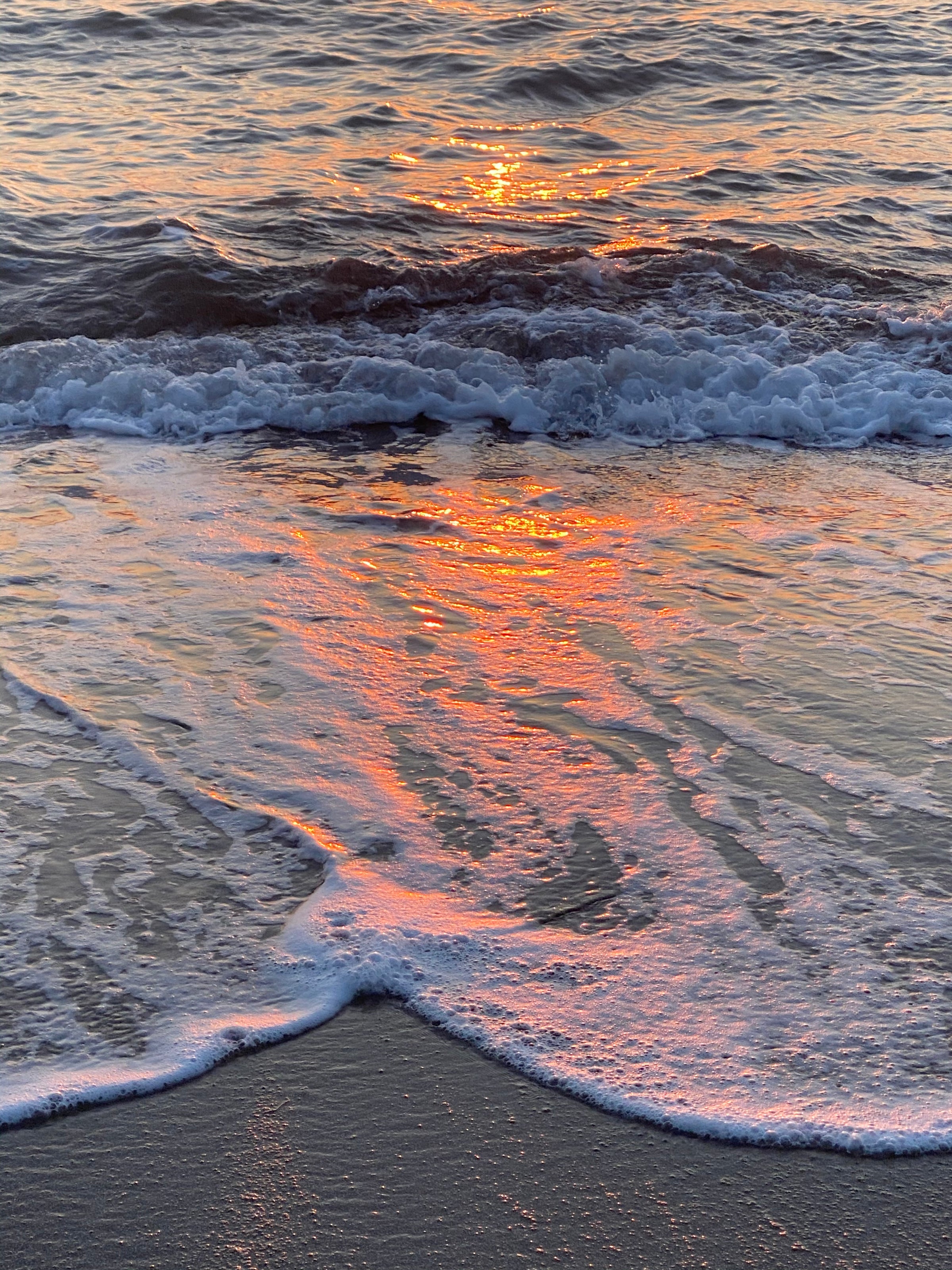 Sunset reflects orange and pink hues on ocean waves as they gently wash onto the sandy shore. The foam creates intricate patterns on the wet sand, and the water shimmers with the glow of the setting sun.