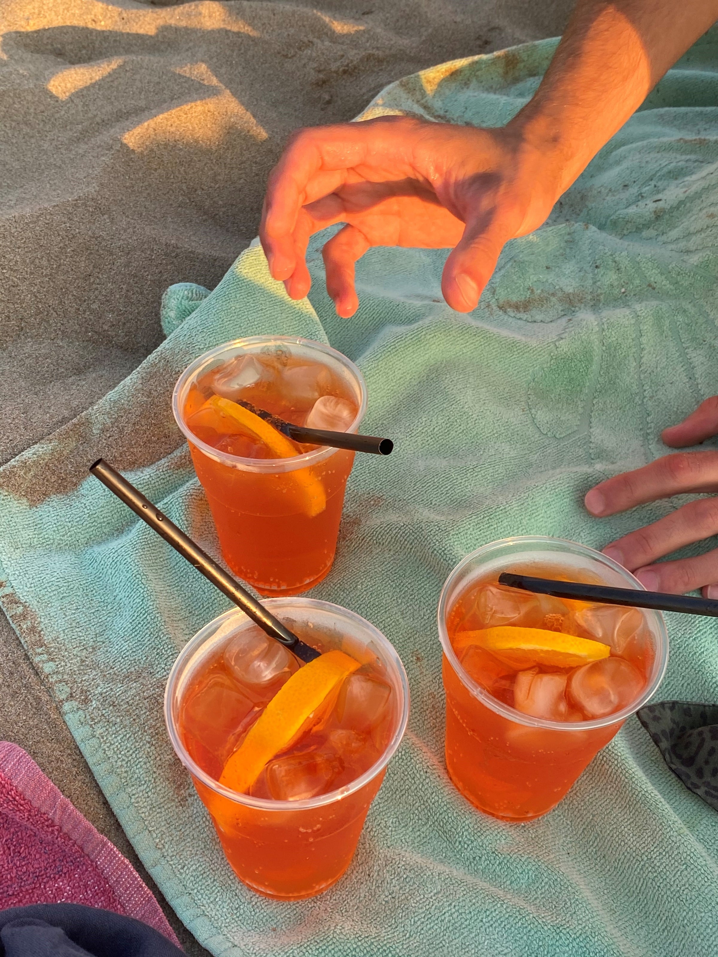 Three plastic cups of orange cocktails with ice and orange slices sit on a beach towel in the sand. Two hands, one reaching and one resting, are near the drinks.