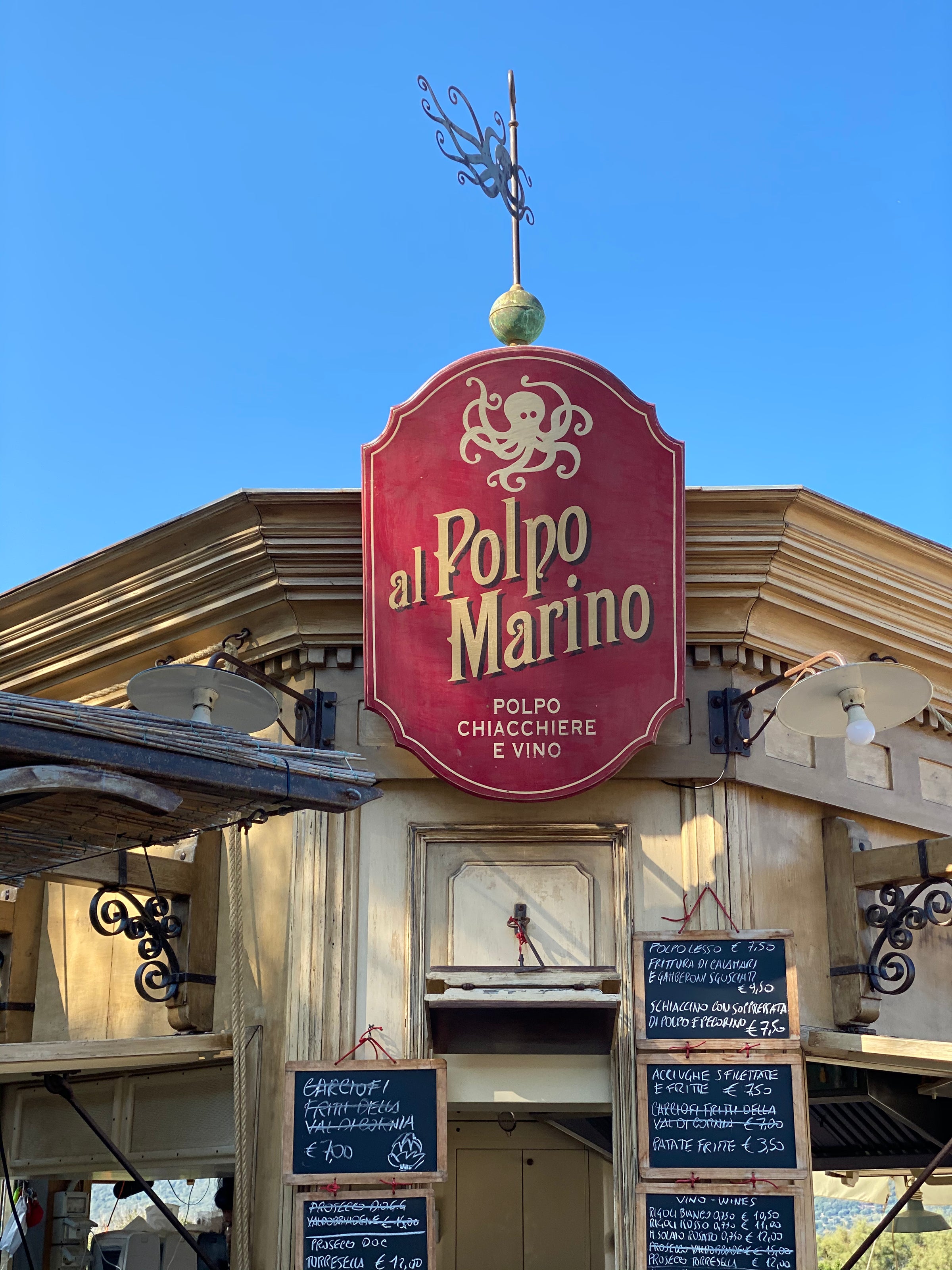A red sign with a gold octopus logo reads al Polpo Marino above the entrance of a rustic seafood restaurant. Several blackboards with handwritten menus are displayed below the sign. Blue sky in the background.