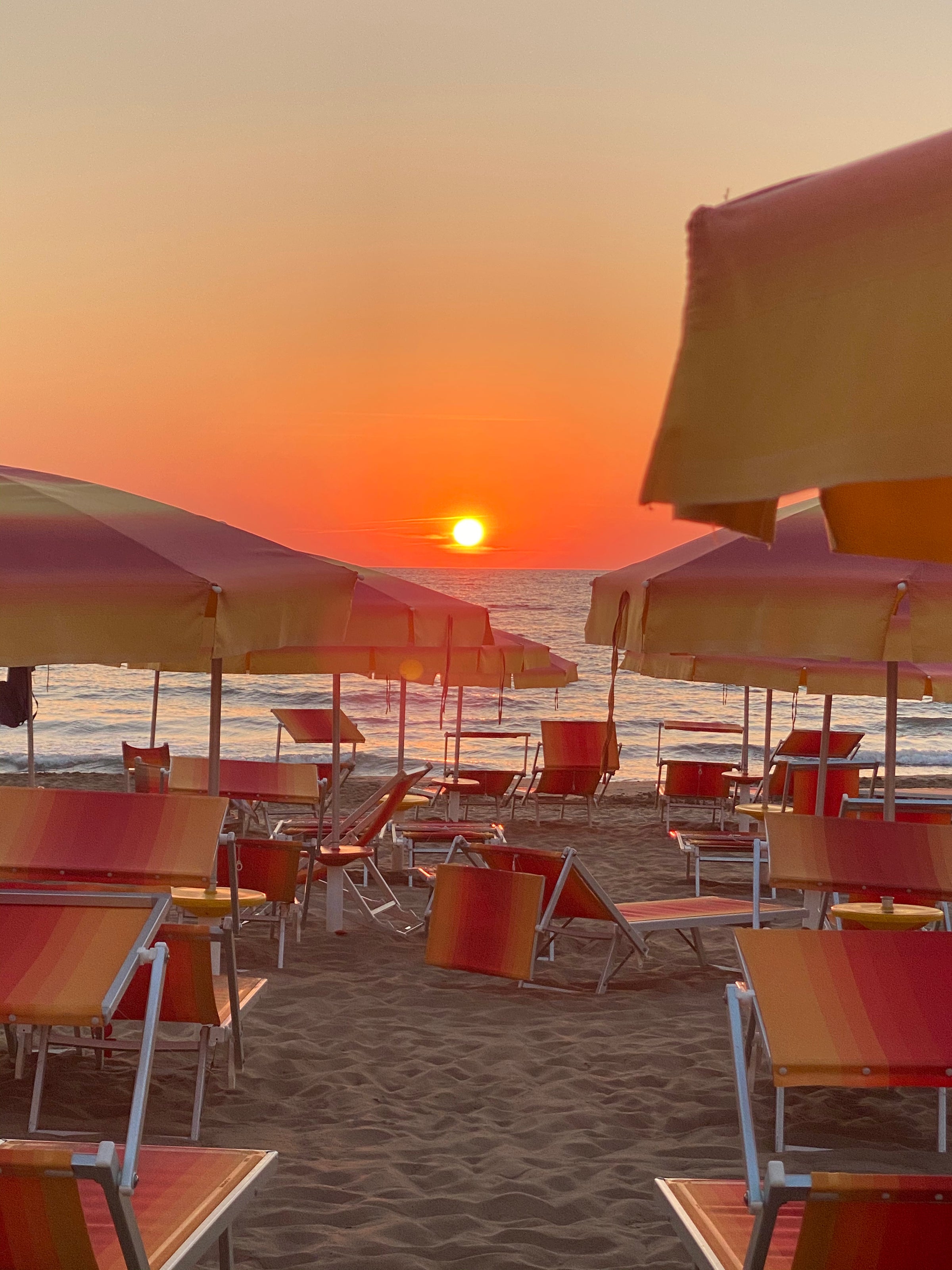 Rows of orange and yellow beach umbrellas and lounge chairs on sandy beach at sunset, with the sun low over the calm sea and the sky glowing in warm orange tones.