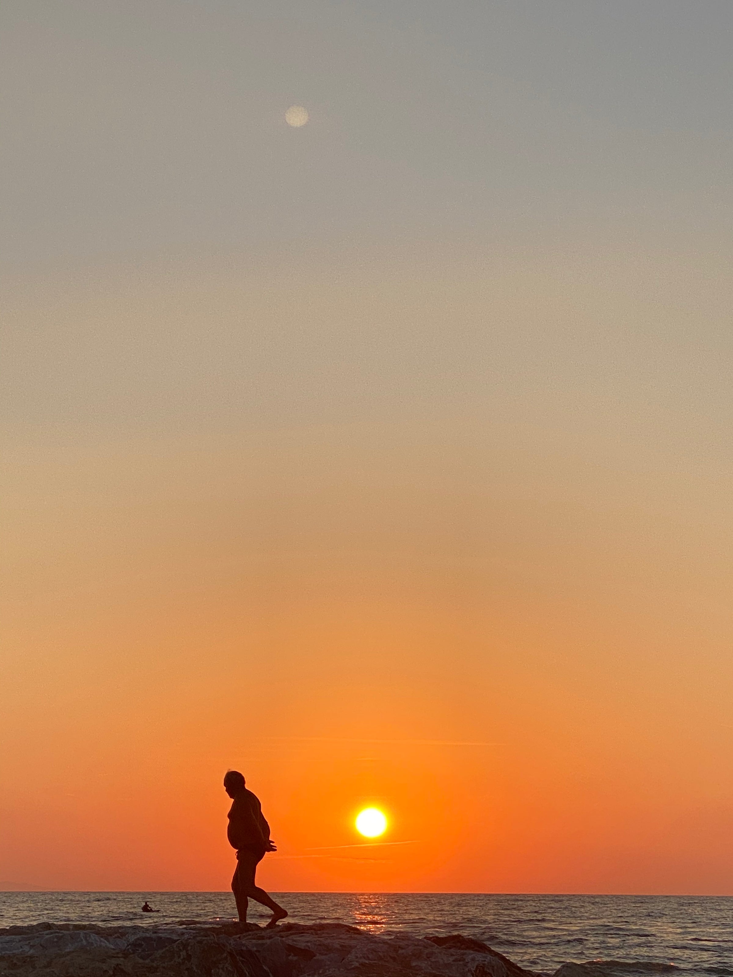 A person is silhouetted against an orange sunset, walking along a rocky shore with the sun low on the horizon, casting a warm glow over the calm sea and sky.
