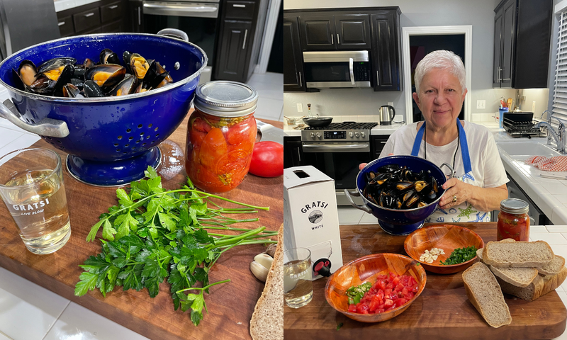 A woman in a kitchen holds a blue colander of cooked mussels. On the counter are fresh parsley, tomatoes, garlic, bread, a jar of tomatoes, a glass of white wine, and prepared bowls of chopped ingredients.