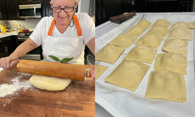A smiling person in an apron rolls out dough with a rolling pin on a wooden surface; on the right, freshly made ravioli rest on a cloth-covered tray in a kitchen.