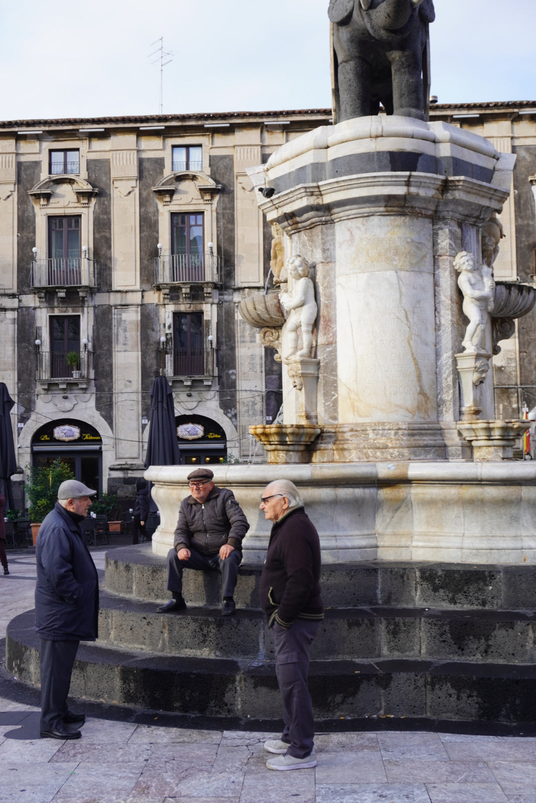 Three elderly men talk near a decorative marble fountain in a European square, with one man sitting and two standing. Behind them is an old, multi-story building with balconies and closed umbrellas.