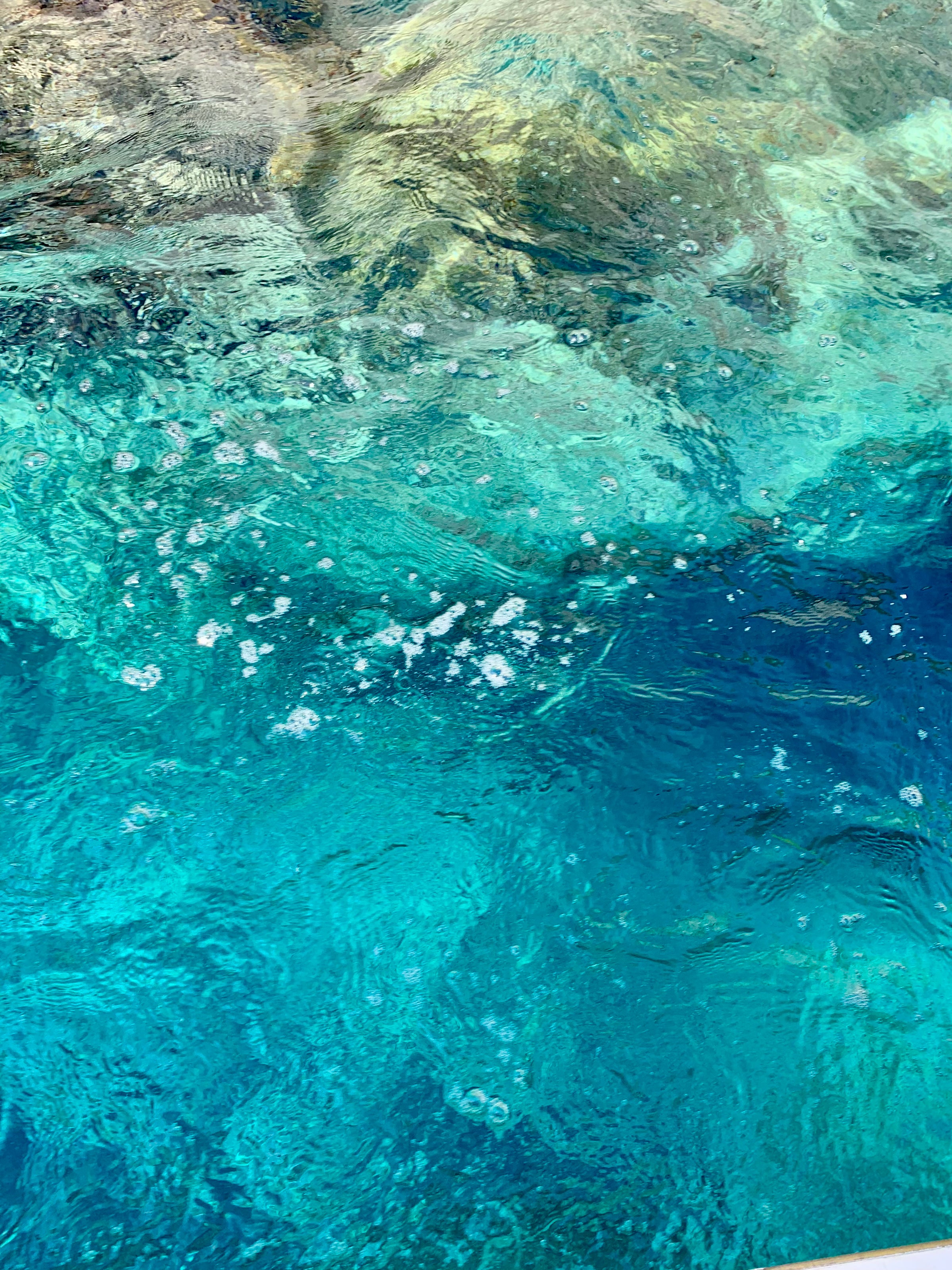 Blue water with visible bubbles and ripples on the surface; lighter and darker shades indicate varying depths and sunlight reflections beneath the water.