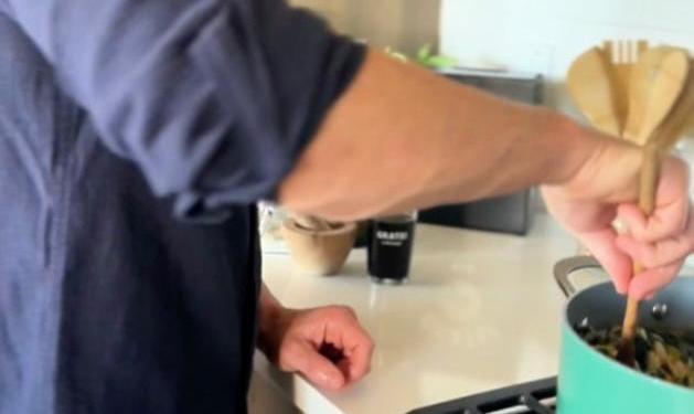 A person in a blue shirt stirs food in a pot on a stove using a wooden spoon. The scene is set in a bright kitchen with a white countertop and various items in the background.