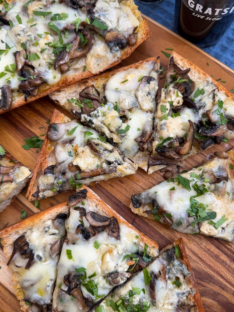 Sliced cheesy mushroom toast topped with chopped parsley on a wooden cutting board, with a partially visible drink on the side.