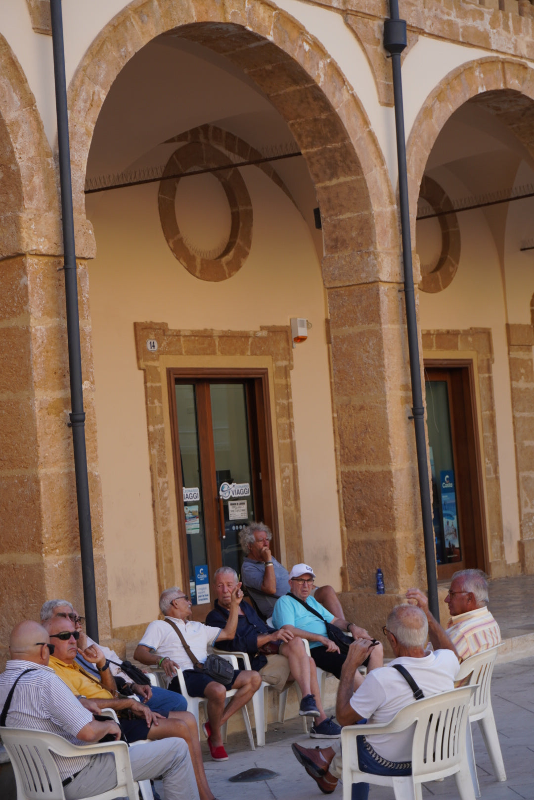 A group of older men sit and chat on white plastic chairs outside a building with stone arches and beige walls, enjoying a sunny day.