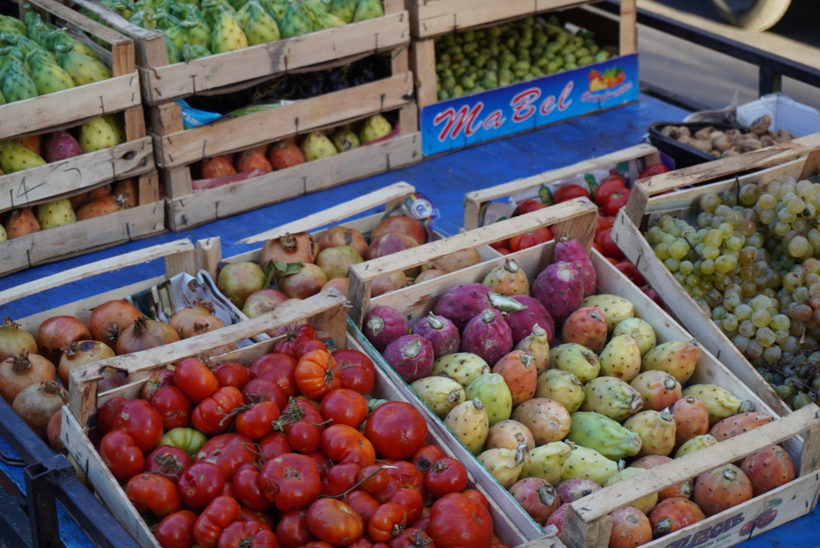 Wooden crates filled with tomatoes, prickly pears, pomegranates, grapes, and other fruits are displayed on a blue table at an outdoor market.