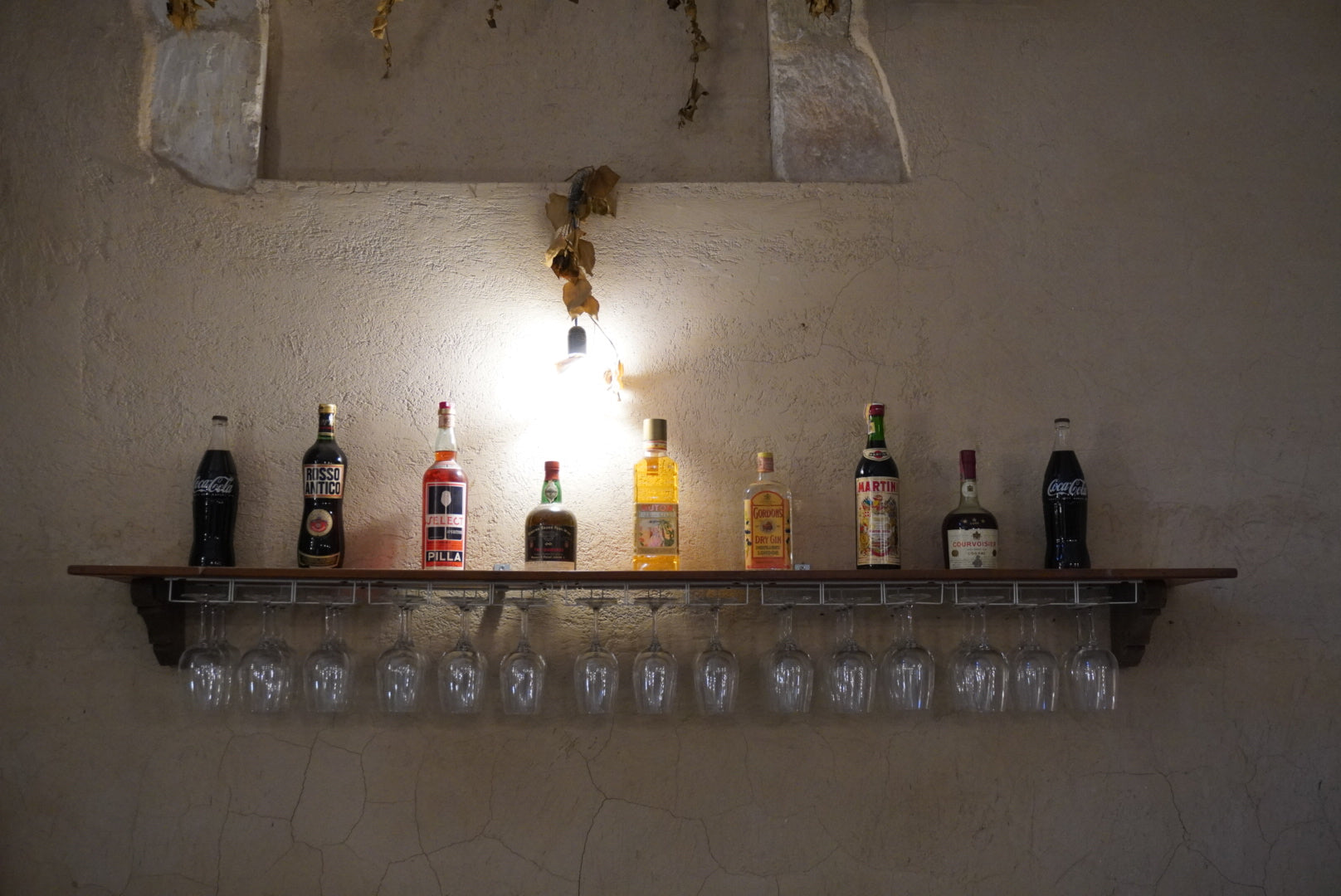A row of liquor bottles and two Coca-Cola bottles sit on a wooden shelf mounted on a textured wall, illuminated by a single exposed bulb above, with wine glasses hanging upside down beneath the shelf.