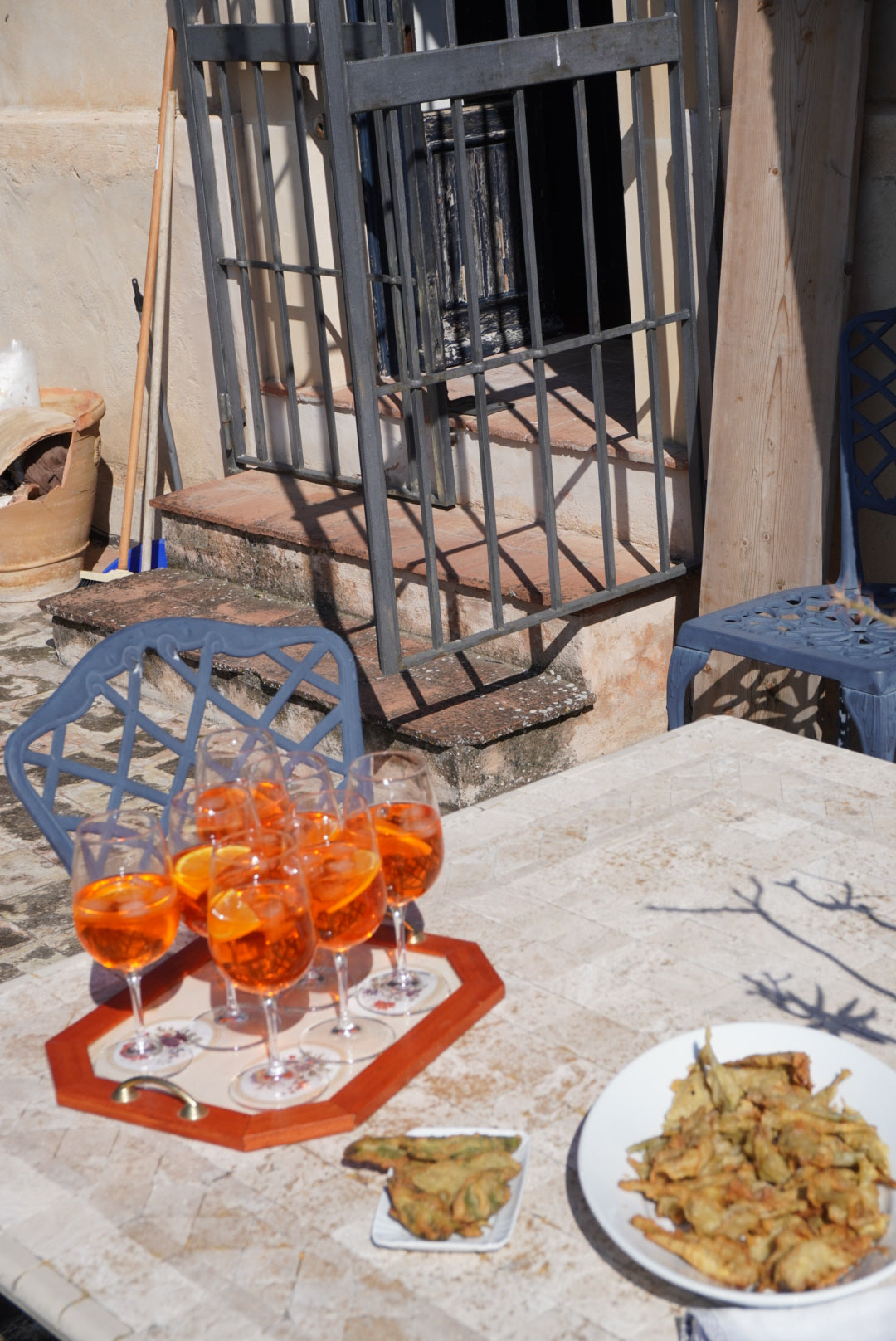 A sunlit outdoor table with six glasses of orange spritz cocktails on a tray and a plate of fried food, possibly vegetables. In the background are metal chairs, stairs, and a gated door.