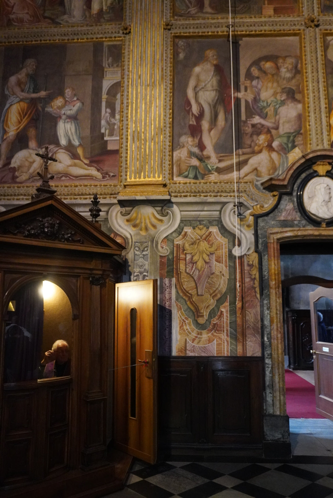 A person sits inside a wooden confessional booth in a richly decorated church with ornate frescoes and colorful paintings covering the walls above.