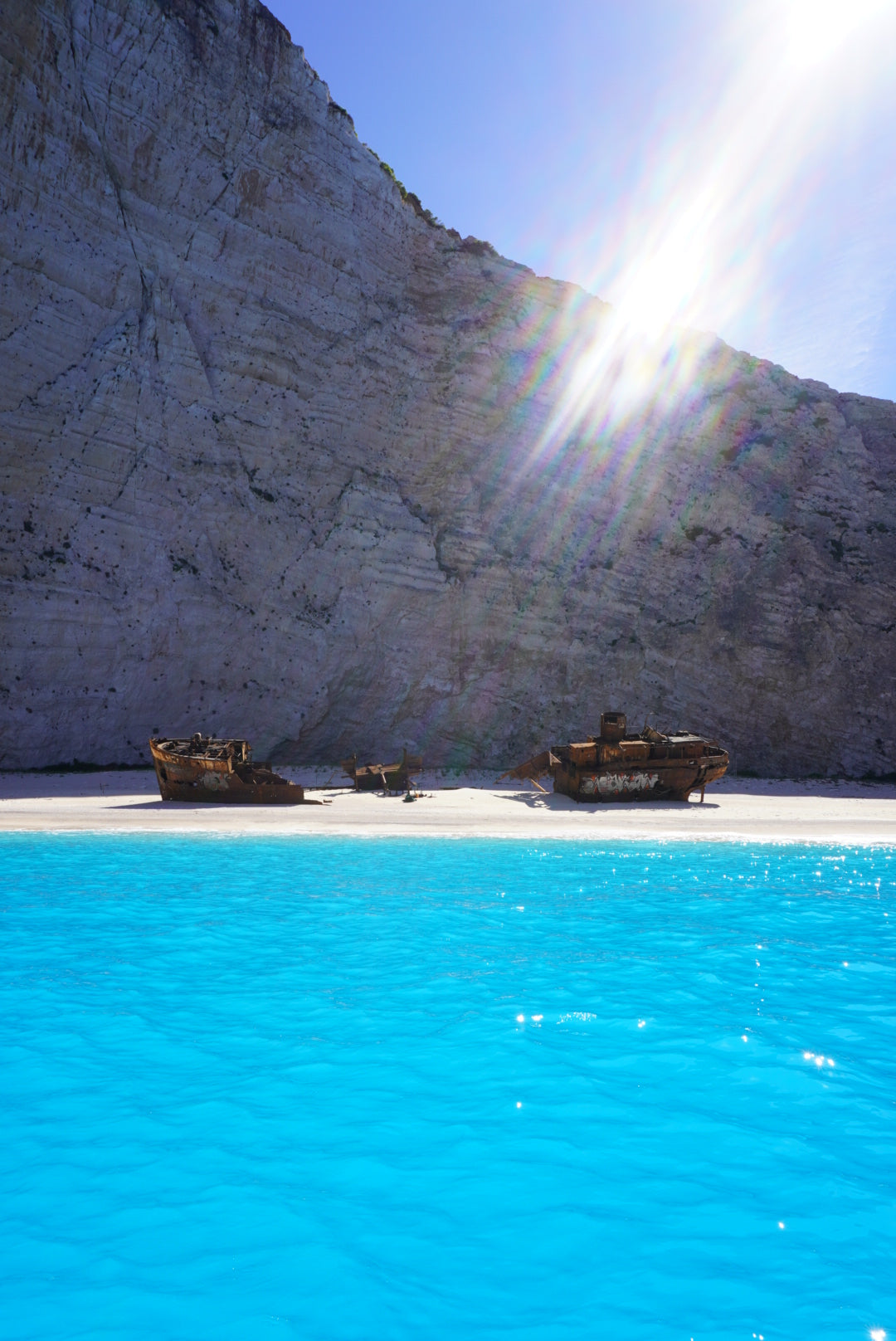 Bright blue water in the foreground, a sandy beach with two rusted shipwrecks in the middle, and steep rocky cliffs behind. Sunlight streams down from the upper right corner of the image.