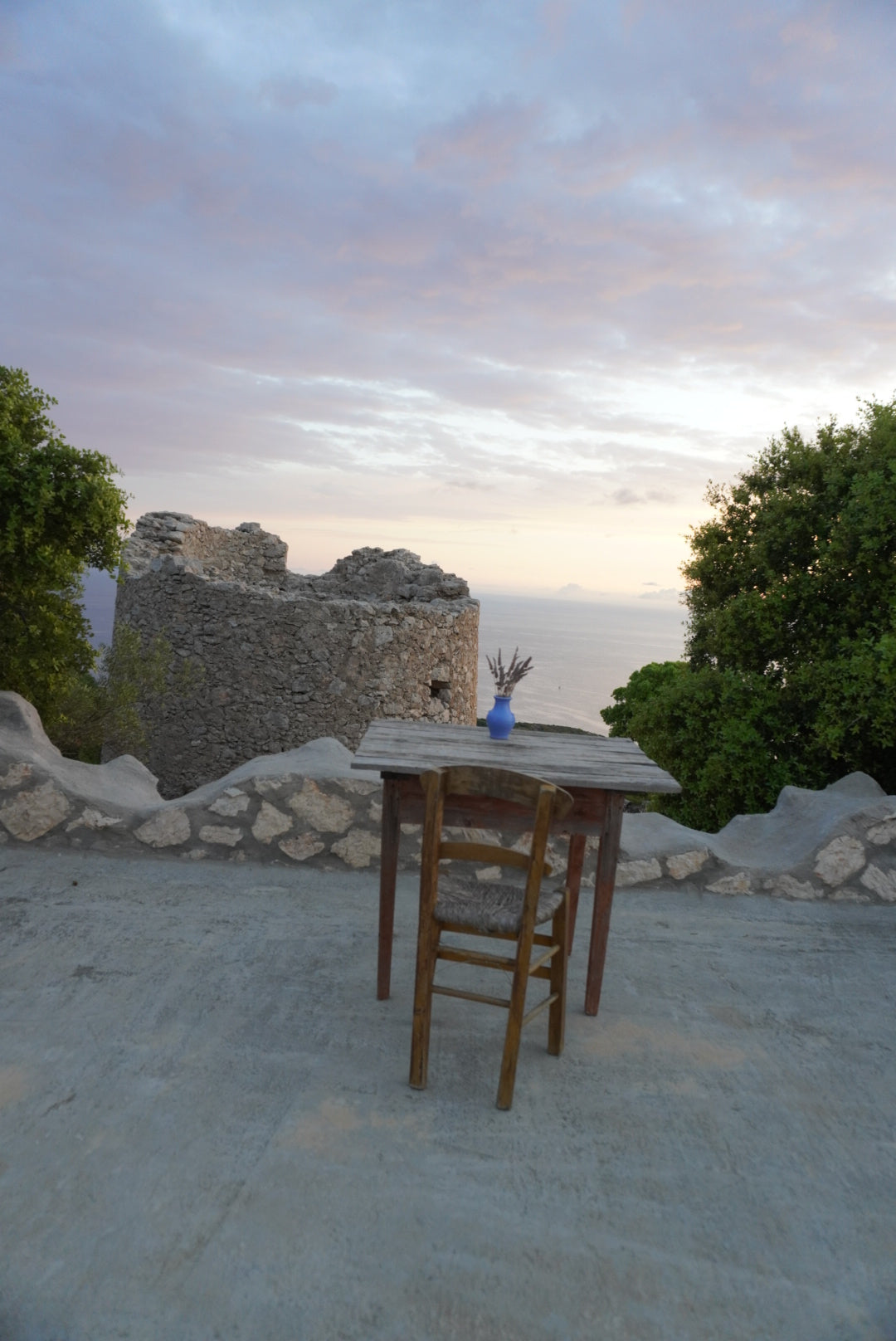 A wooden chair and table with a blue vase sit on a stone patio overlooking an old stone ruin, lush green trees, and a calm sea at sunset under a partly cloudy sky.