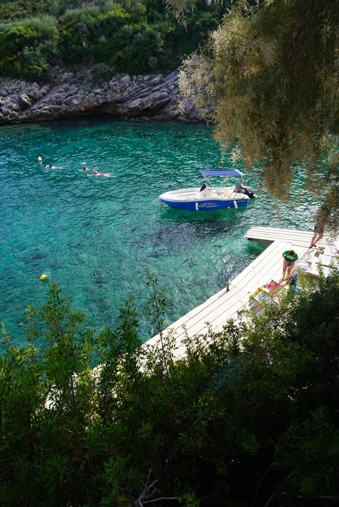 A small motorboat floats near a wooden dock on clear turquoise water, with people swimming and sunbathing nearby. Lush green trees and rocky shoreline frame the peaceful scene.