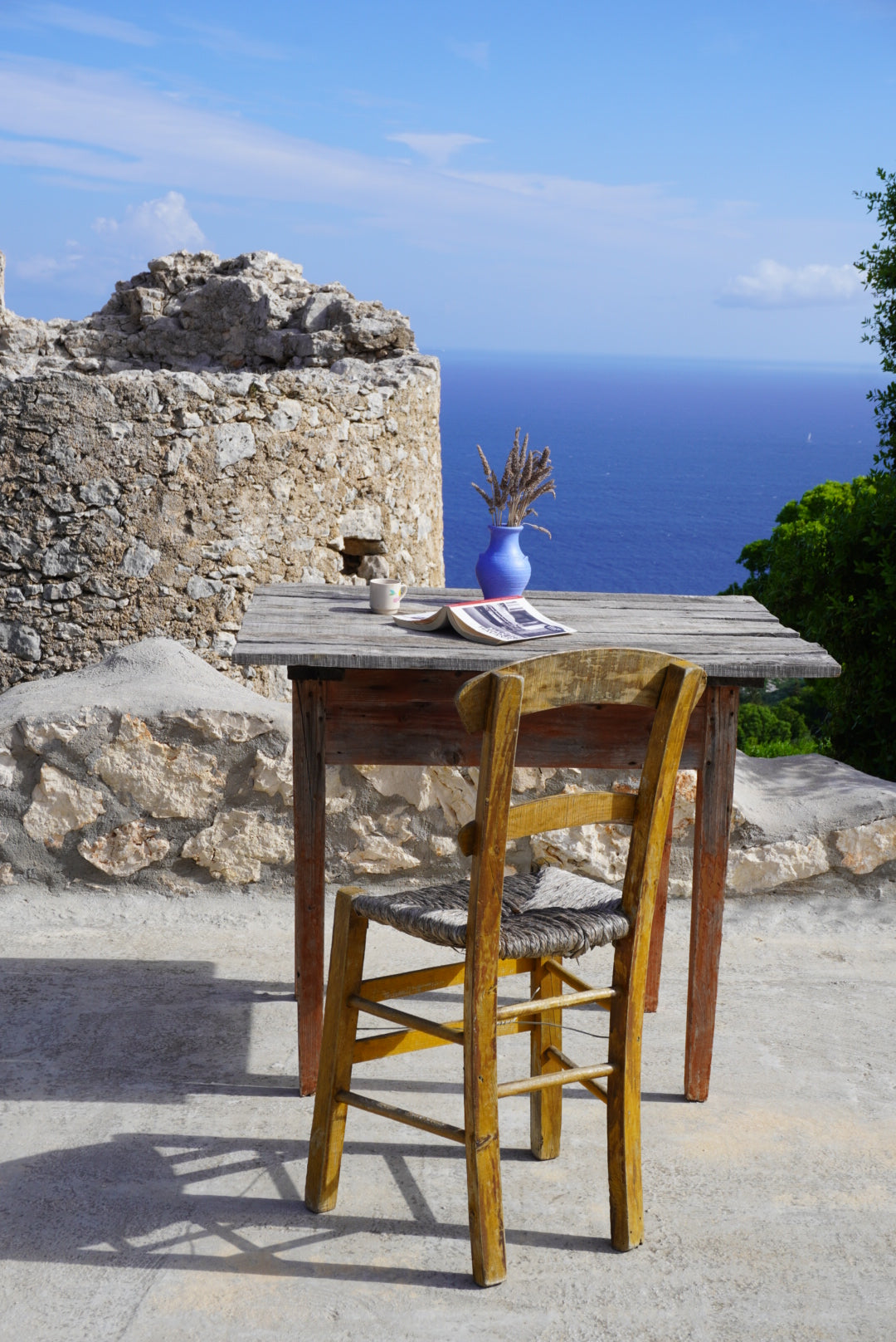 A rustic wooden table and chair sit on a stone terrace overlooking the sea. On the table are a blue vase with dried flowers, a cup, and papers. In the background are old stone ruins and a clear blue sky.