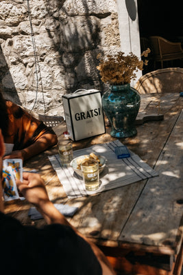 Two people sit at a rustic wooden table outdoors, playing cards. On the table are drinks, a box labeled GRATSI, dried flowers in a vase, and sunlight casting shadows through nearby foliage.