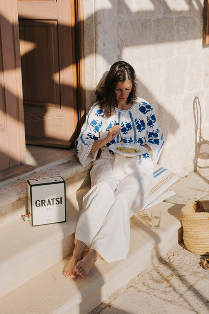 A woman in white pants and a blue-and-white embroidered blouse sits on stone steps in sunlight, eating from a bowl. Beside her is a box labeled “GRATSI,” a glass of white wine, and a woven bag.