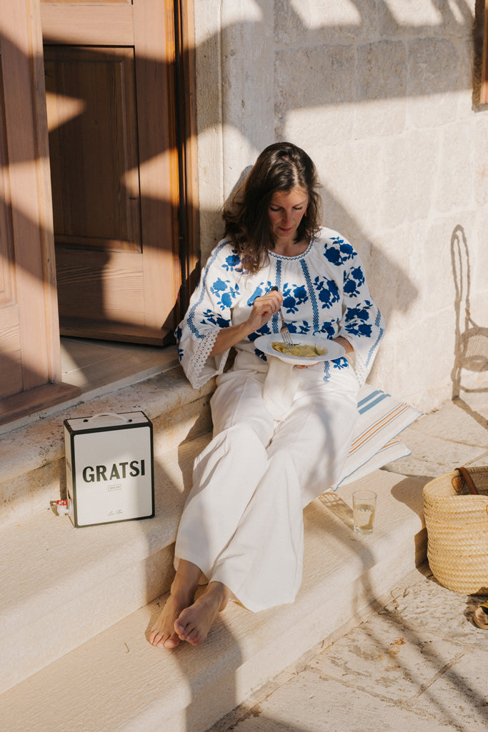 A woman in white pants and a blue-and-white embroidered blouse sits on stone steps in sunlight, eating from a bowl. Beside her is a box labeled “GRATSI,” a glass of white wine, and a woven bag.