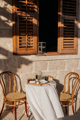 A small round table with a white tablecloth is set for two with food, drinks, and flowers, placed outside against a stone wall and beneath an open wooden window. Two wooden chairs are positioned on either side of the table.