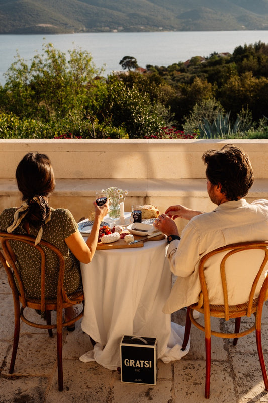 Two people sit at a small outdoor table with wine and snacks, overlooking a scenic view of trees, hills, and water. The scene is warm and inviting, with sunlight casting a cozy glow on the terrace.
