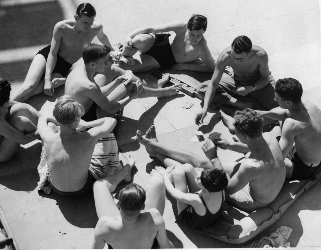 A group of young men in swim trunks sit in a circle on a sunny concrete surface, playing cards and talking. Towels and playing cards are visible on the ground.