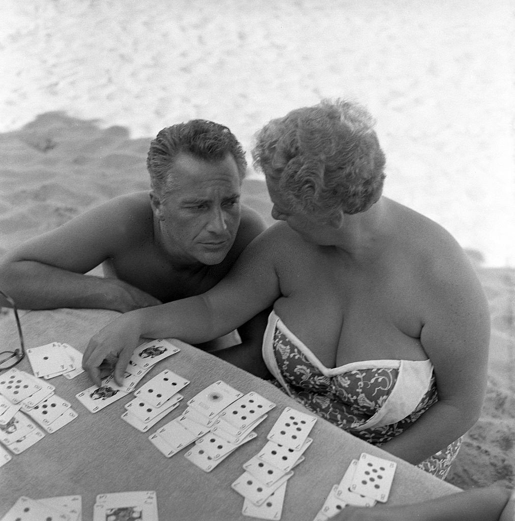A man and a woman in swimsuits sit at a table on the beach, playing cards. The woman is arranging playing cards on the table, while the man leans in, looking at her and the game. Sand is visible in the background.