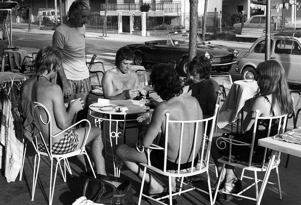 Six people, mostly shirtless and in swimwear, sit around a small outdoor table at a café or patio, playing cards. Vintage cars and buildings are visible in the background. The photo is black and white.