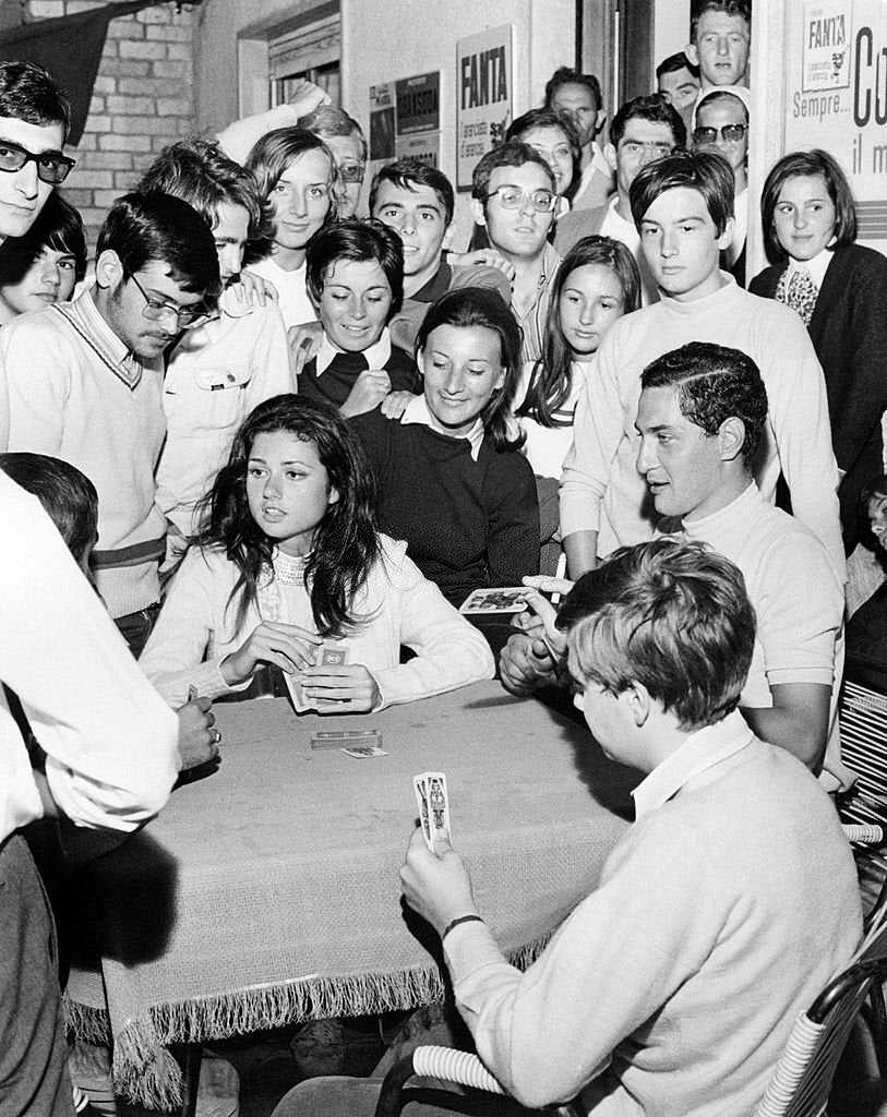 A group of young people gathered around a table playing cards, with many others standing and watching closely. The atmosphere appears lively and engaging, suggesting a friendly social gathering.