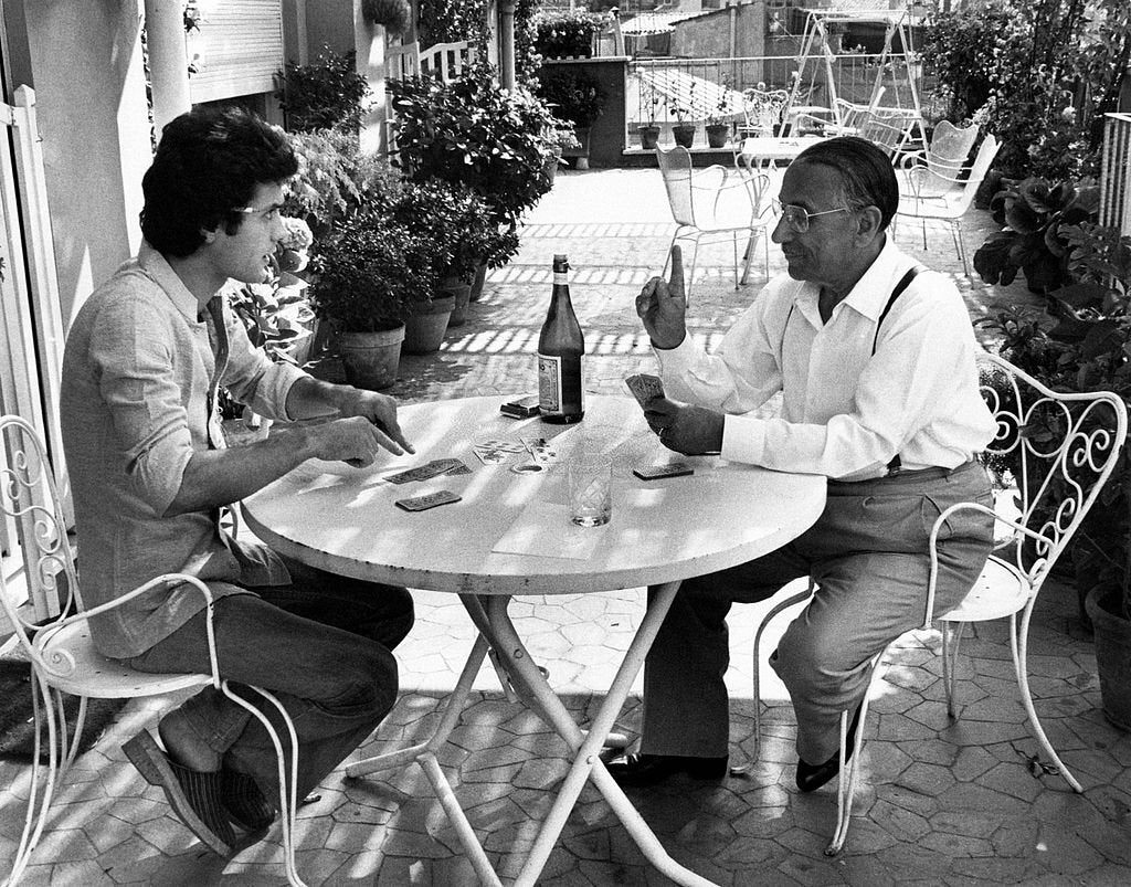 Two men sit at a round table outdoors, playing cards. Potted plants surround them, and drinks and a bottle are on the table. One man gestures with his hand, while the other looks at his cards. The setting is bright and relaxed.