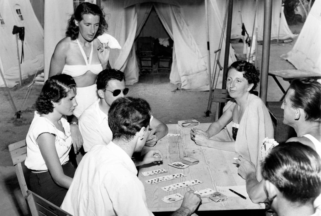 Six people sit around a table playing cards outside near large tents; one woman stands behind, watching the game, while others sit and focus on the cards. The scene appears relaxed and informal.
