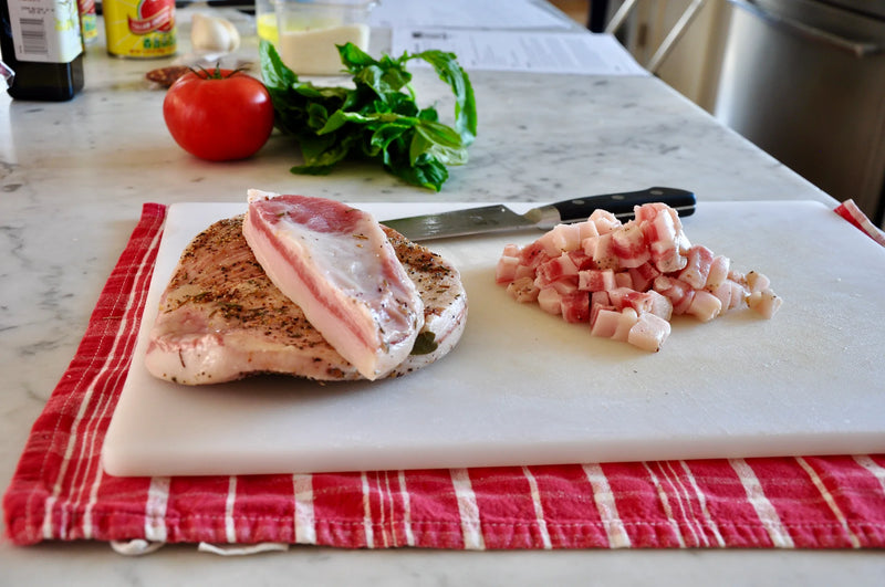 A slab of seasoned pork and diced pork pieces sit on a white cutting board with a knife. Fresh basil and a tomato are in the background on a marble countertop. The board rests on a red kitchen towel.