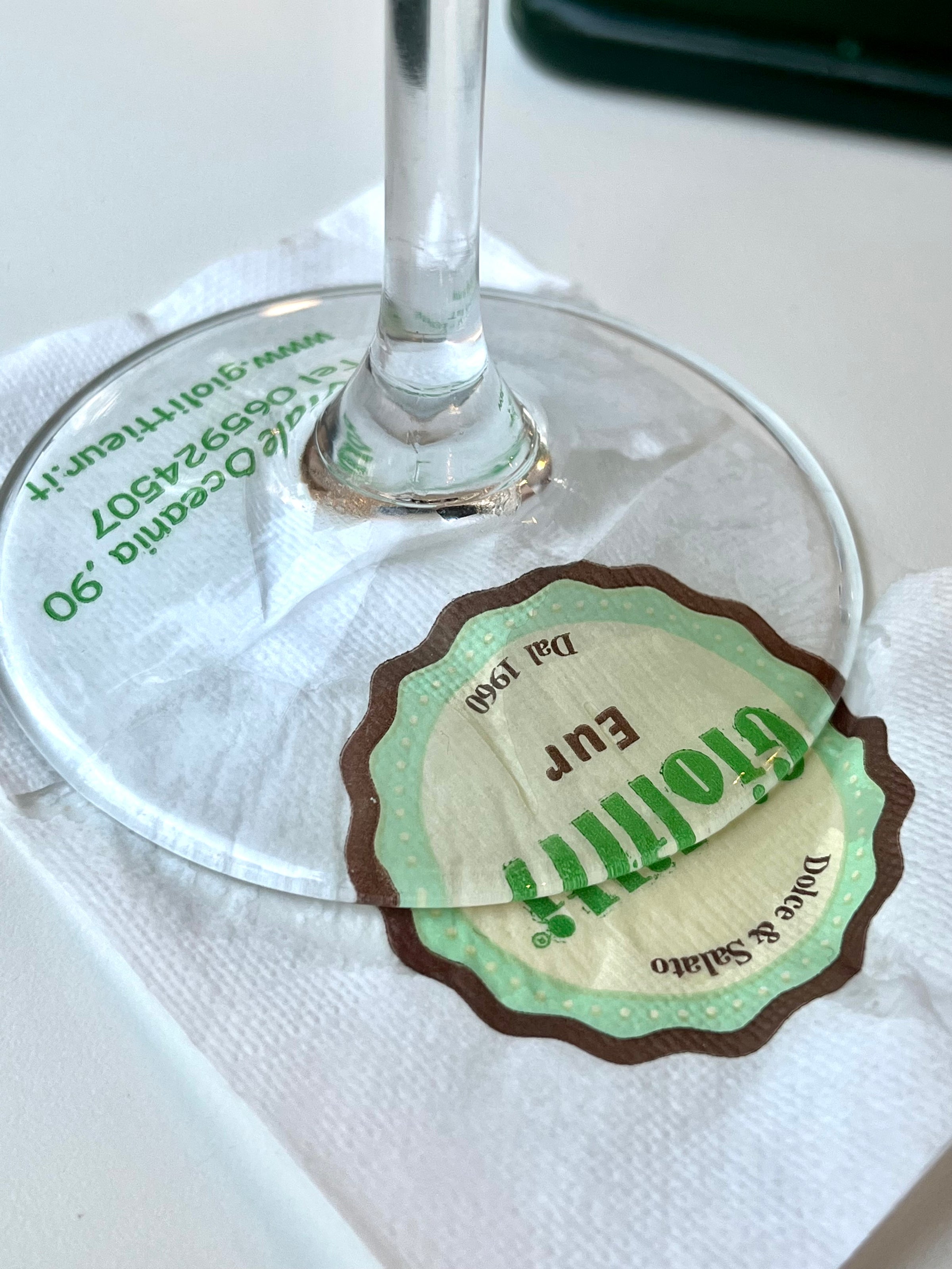 A wine glass rests on a white napkin, with a green and brown sticker labeled Grolift! EUR and some small printed text in Italian visible through the glass base.