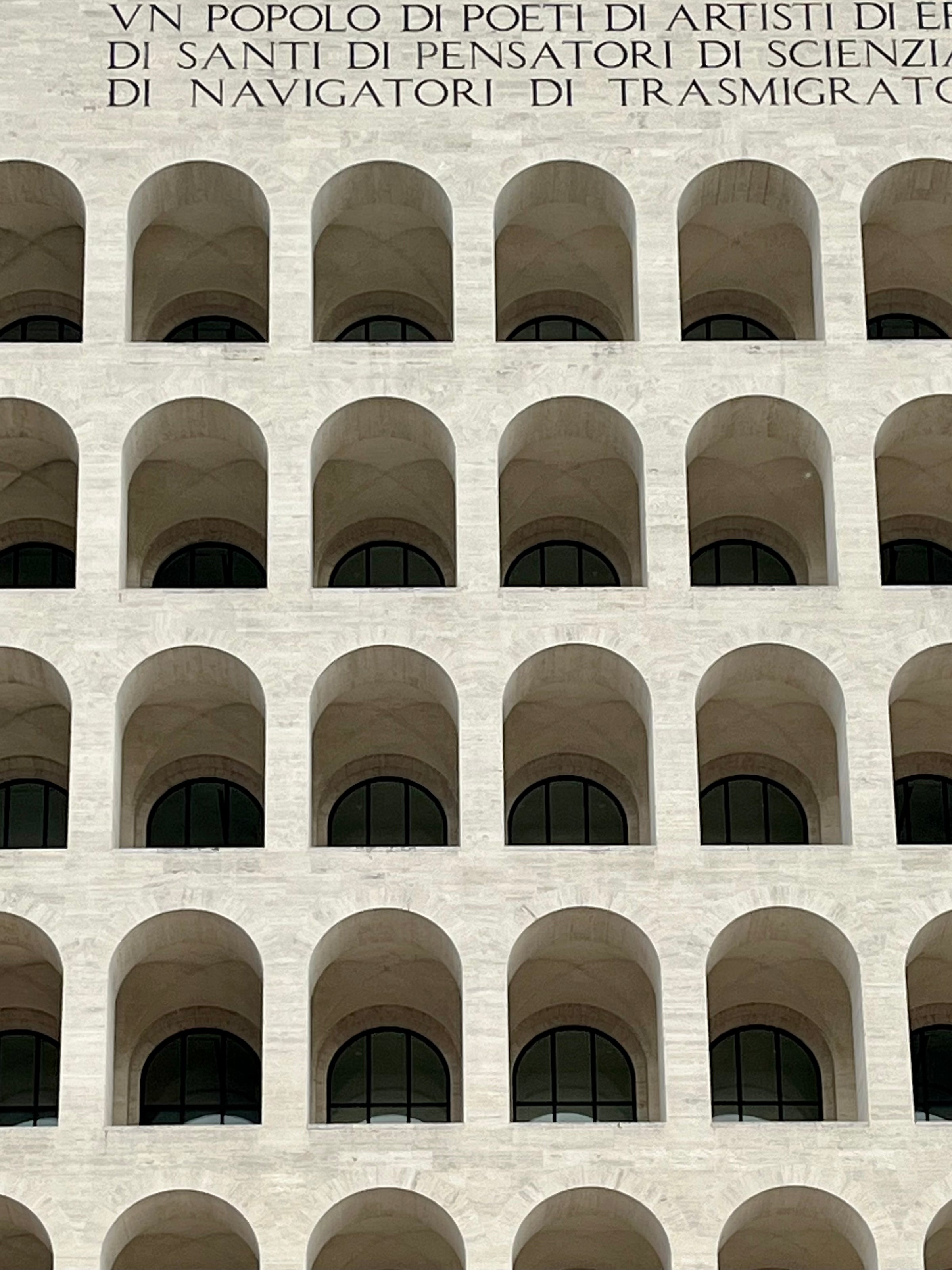 A close-up of a white stone building facade featuring multiple rows of evenly spaced arched windows. Above the arches, Italian text is engraved into the stone.
