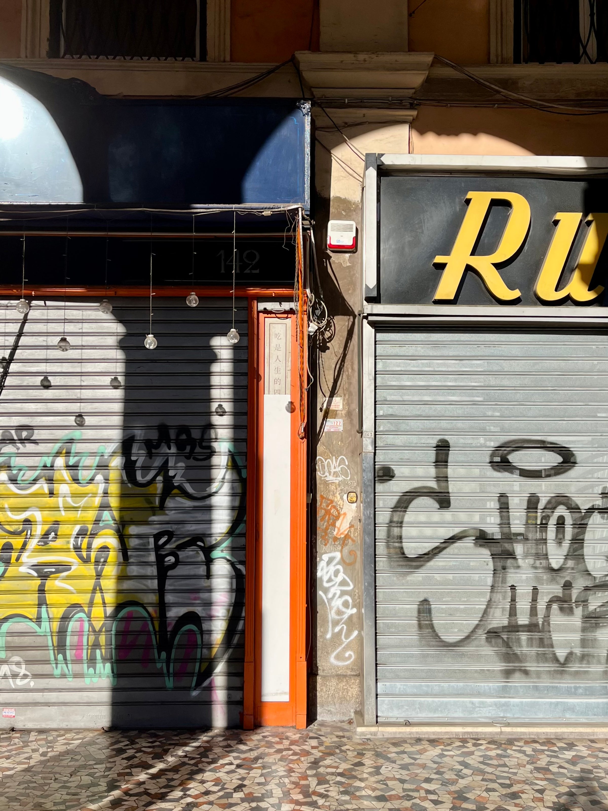 Two closed metal shop shutters, both covered in graffiti, are shown in bright sunlight. The shop on the left has a dark blue sign, while the one on the right has a black sign with yellow letters partially visible.