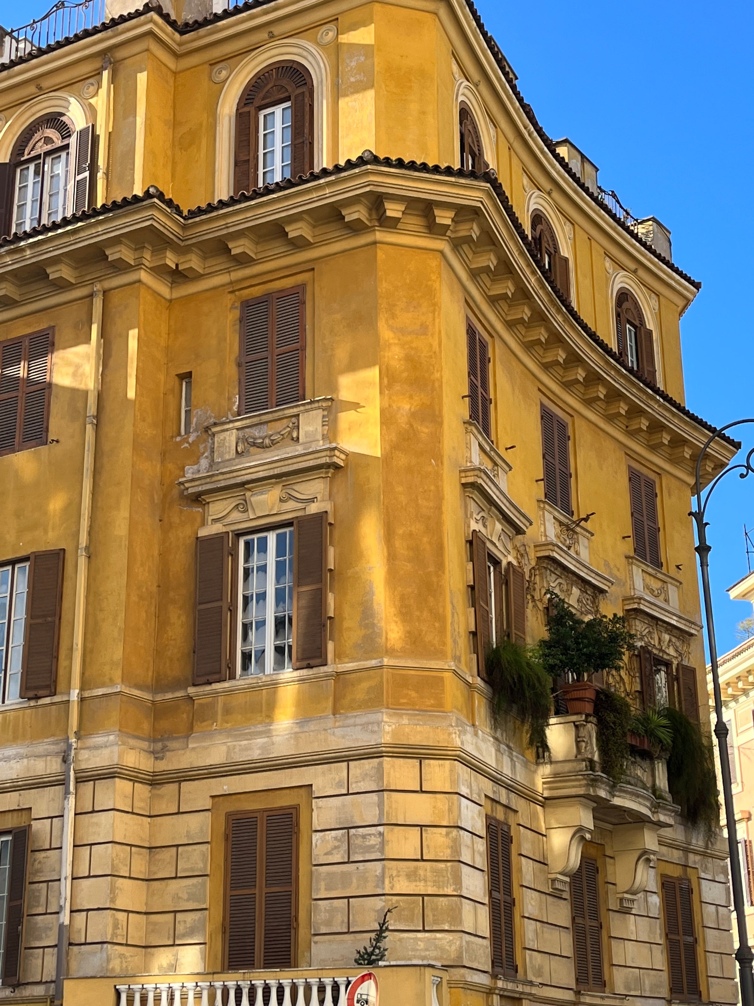 A yellow, historic European building with arched windows, wooden shutters, balconies with plants, and ornate architectural details, set against a clear blue sky.