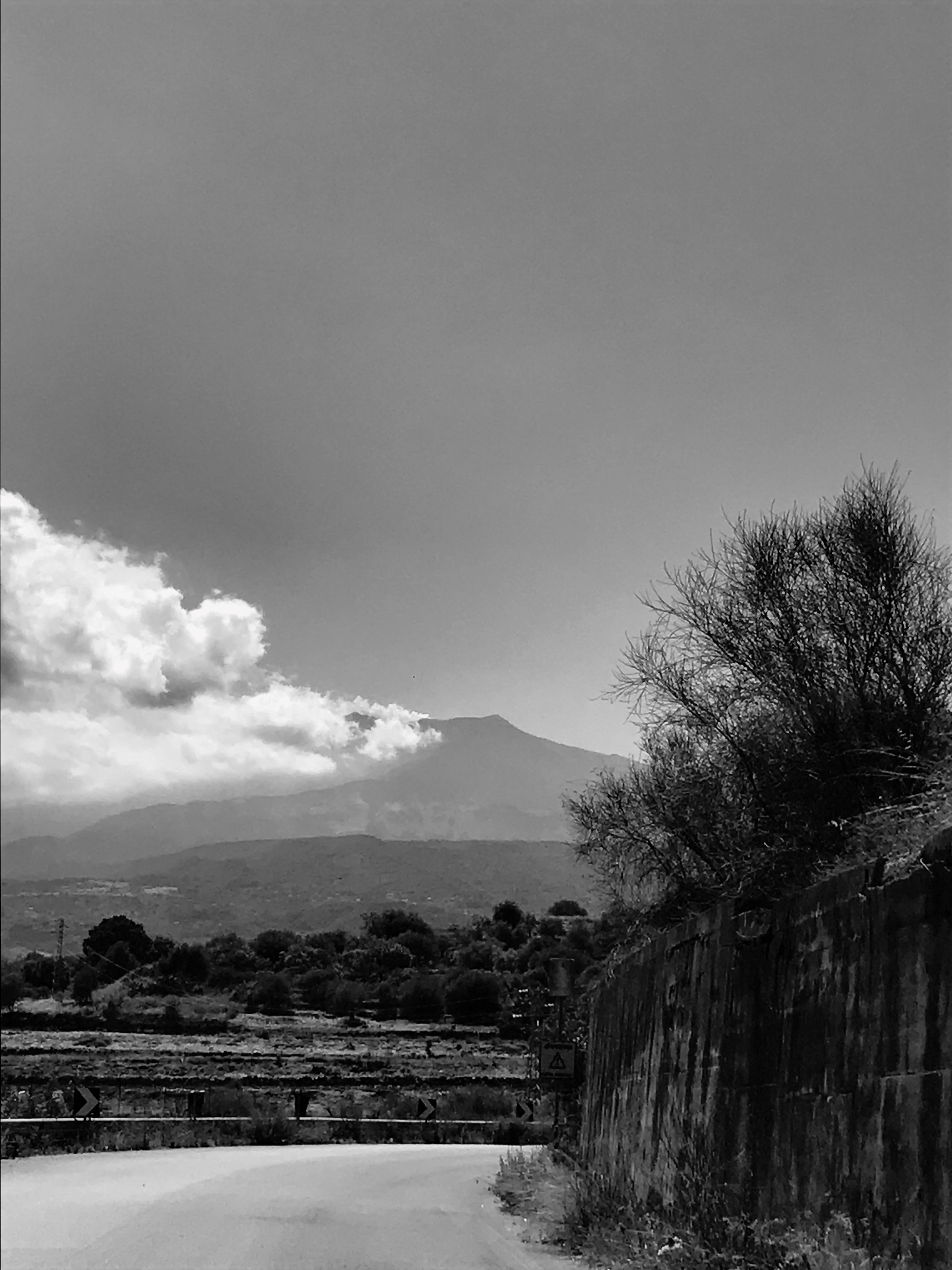 A black and white photo of a rural road curving beside a stone wall, with dry bushes and trees. In the background, a mountain with smoke or clouds near its peak is visible under a hazy sky.