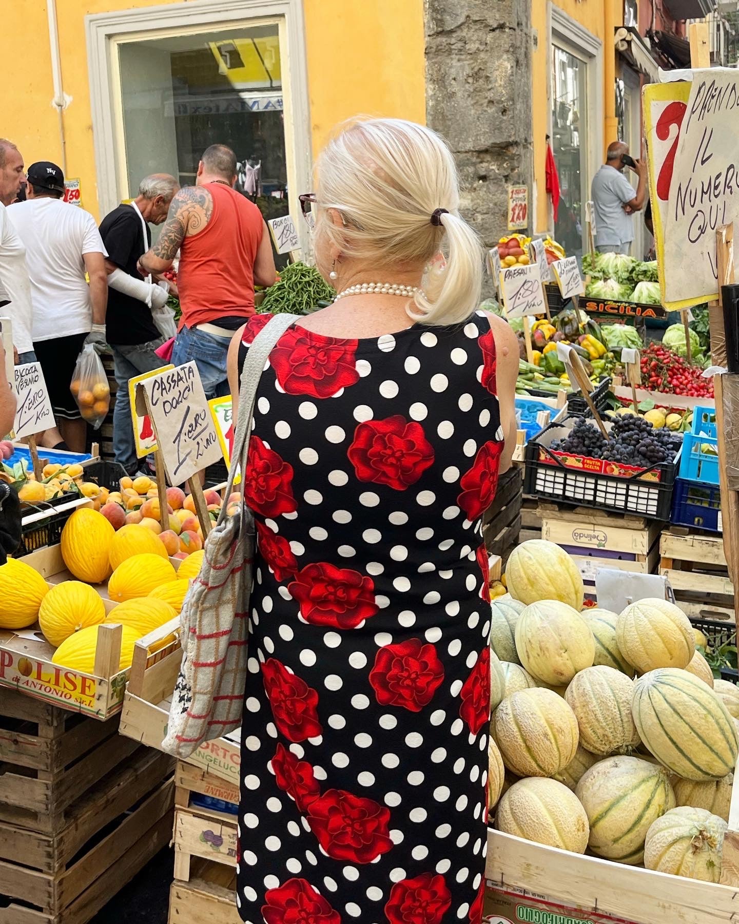 An older woman with white hair in a ponytail, wearing a black dress with white polka dots and red roses, stands at an outdoor fruit and vegetable market. Signs, crates of melons, and other shoppers are visible around her.
