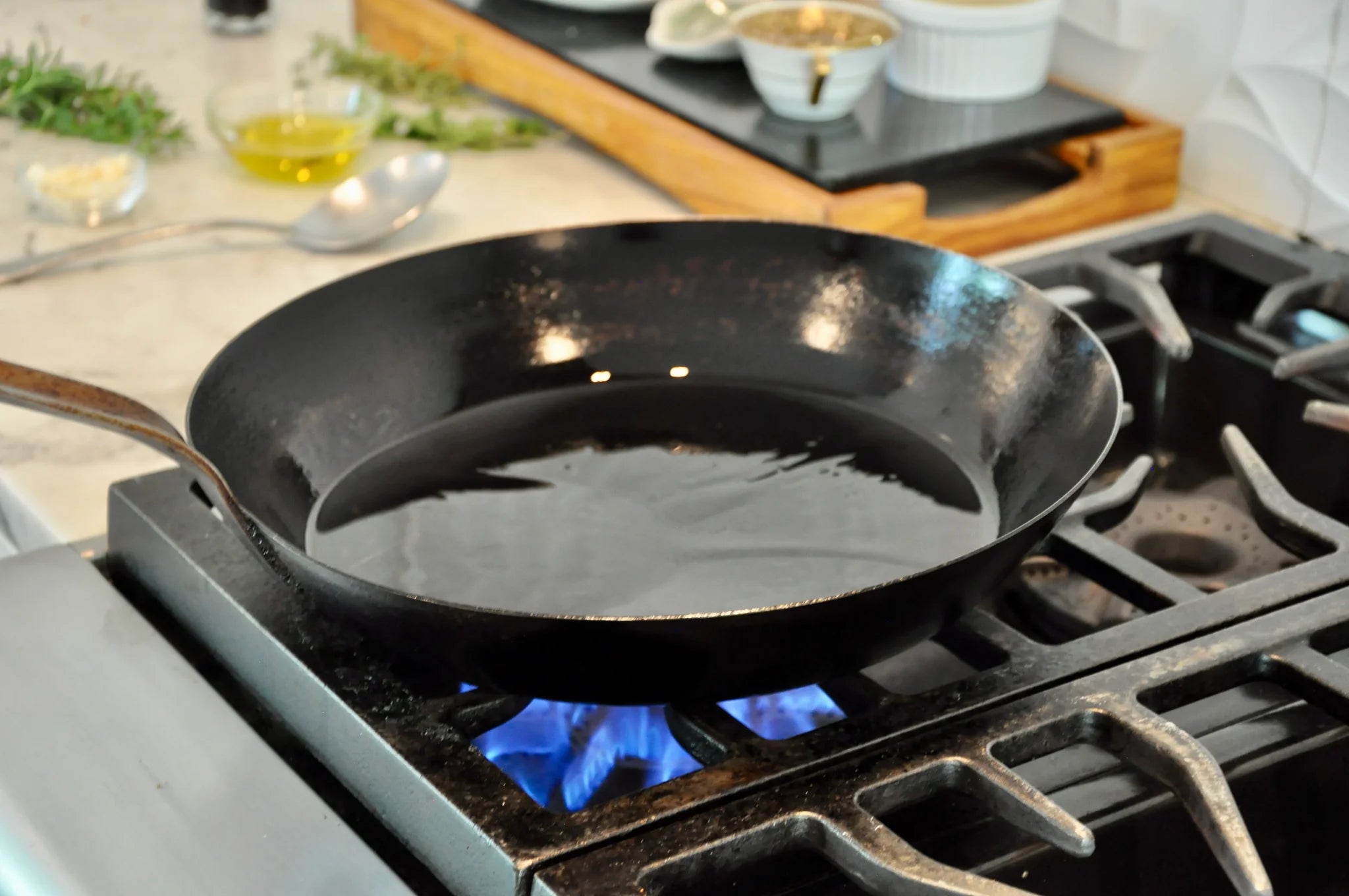 A cast iron skillet with oil is heating on a gas stove with blue flames, in a kitchen setting with various ingredients and utensils in the background.
