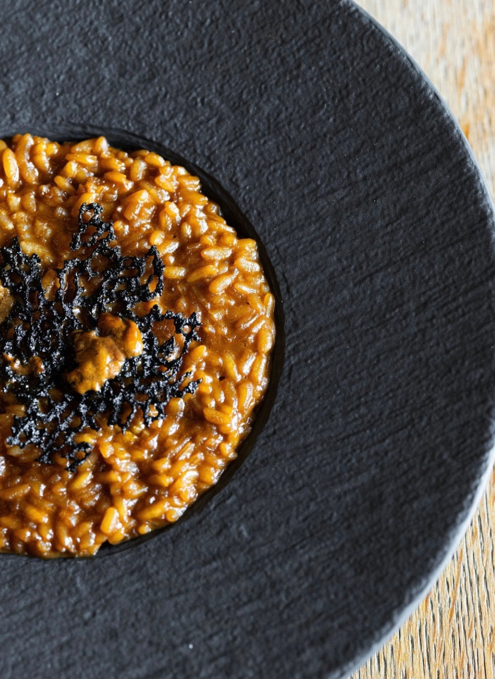 A close-up of a plate of creamy risotto with a rich brown sauce, garnished with a decorative black lace-like crisp on top, served on a textured black plate.