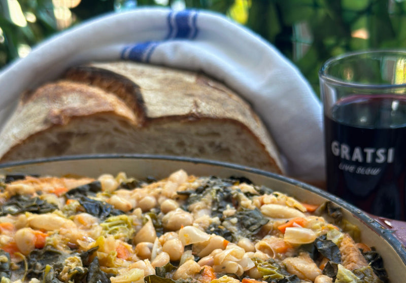 A close-up of a hearty vegetable and bean casserole, with a loaf of rustic bread and a glass of red wine labeled GRATSI set against a leafy green background.