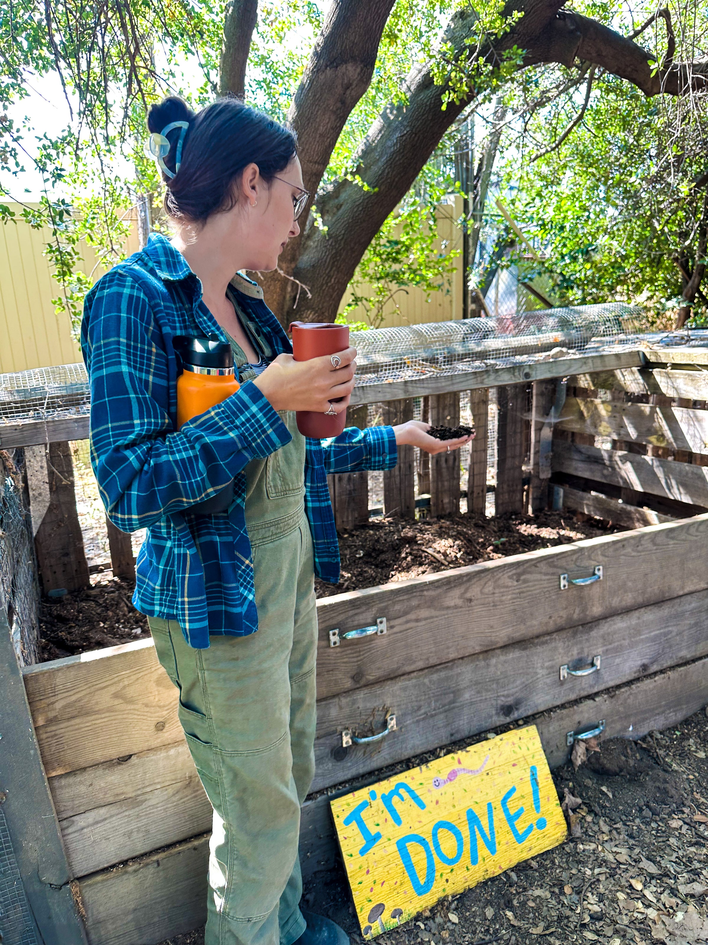 A person in overalls and a plaid shirt stands by a compost bin, holding a travel mug and showing something in their hand. A colorful sign on the bin reads IM DONE! Trees and a fence are in the background.